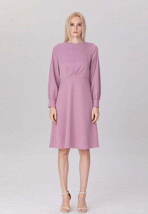Femme portant une robe rose poudré mi-longue à manches longues et des escarpins blancs à bride cheville, debout devant un fond clair uni.