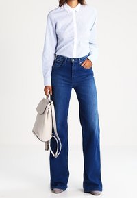 Striped blue button-up shirt paired with blue flared jeans. Model holding a light grey shoulder bag. Soft texture and tailored fit.