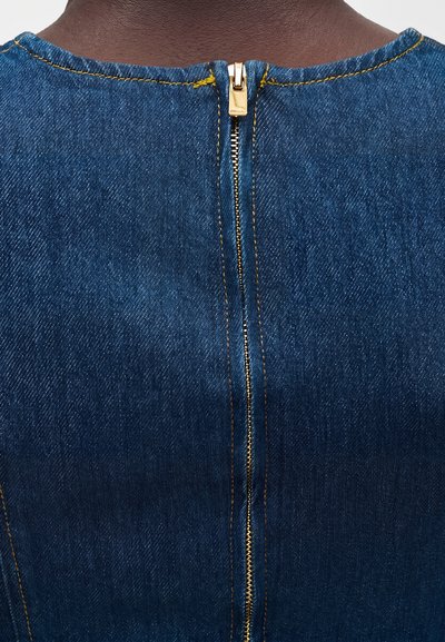 Πίσω μέρος του λαιμού ενός ατόμου με σκούρο μπλε ενδυμασία από τζιν, με ορατό χρυσό φερμουάρ και αντίθετη κίτρινη ραφή.