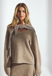 Beige tröja i mjukt stickat material, med avslappnad passform, bred halsringning och långa, lösa ärmar, med en knuten grå scarf runt halsen.