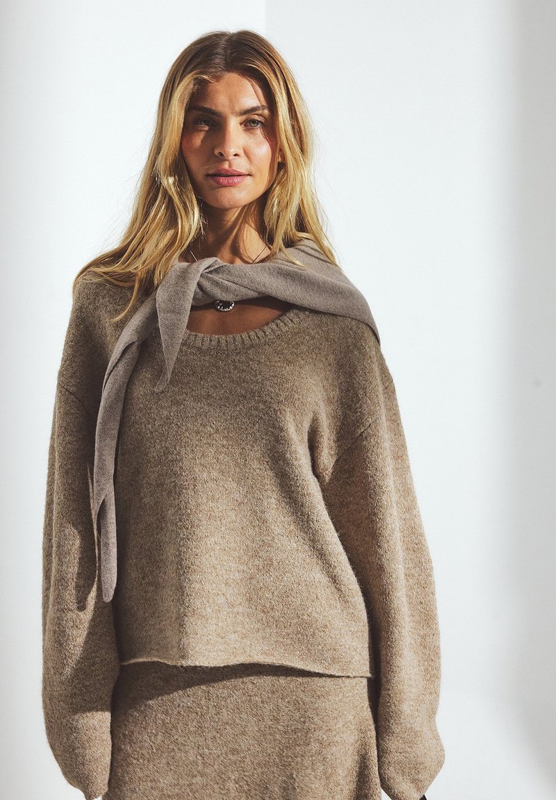 Beige-neule, joka on valmistettu pehmeästä kudotusta materiaalista. Siinä on rento istuvuus, leveä kaula-aukko ja pitkät, löysät hihat, joiden ympärille on solmittu harmaa huivi.