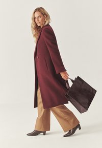 Abrigo burdeos con un diseño entallado, bolso tote de cuero marrón oscuro, pantalones de pierna ancha color beige y zapatos de tacón puntiagudos marrones. Textura suave en todo.