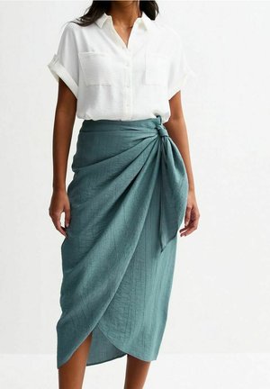 Femme portant une chemise blanche à manches courtes boutonnée et une jupe portefeuille texturée couleur sarcelle nouée à la taille.