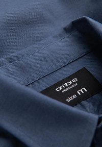 Colletto di camicia blu scuro in tessuto testurizzato. L'etichetta riporta "ombre menswear", indicando la taglia media. Dettaglio in primo piano mostra la cucitura.