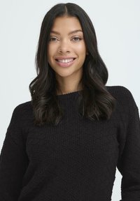 Ung kvinde med langt mørkt hår, som smiler, iført en struktureret sort sweater mod en enkel lys baggrund.
