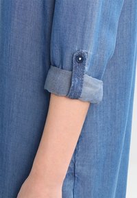 Ljusblå denimskjorta med rullad ärm och knappdetalj. Tyget är mjukt med en slät yta och subtila nyanser.