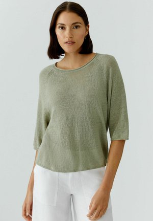 Jonge vrouw met een lichtgroene gebreide trui met driekwart mouwen en een witte broek, staand tegen een effen lichte achtergrond.