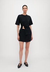 Svart bomull t-skjorte med knute detalj i sentrum. Kombinert med et svart skjørt. Modellen har på seg svarte netting sko. Minimalistisk design.