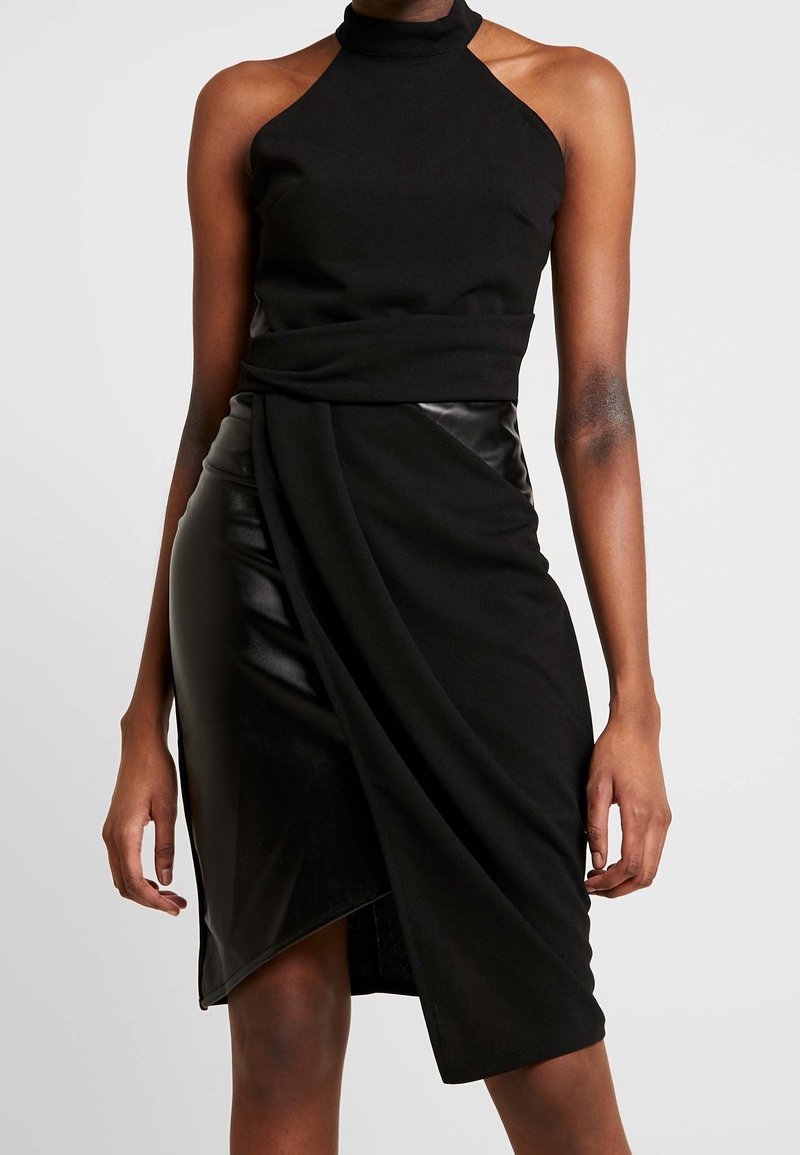 Robe noire sans manches à col haut, jupe portefeuille asymétrique en cuir et tissu, portée par une personne debout sur un fond uni.