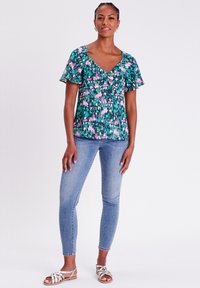 Blumenbluse mit grünem Grundton und pinken sowie lila Mustern, kurzen Schmetterlingsärmeln und einem V-Ausschnitt. Kombiniert mit hellblauen Skinny-Jeans.