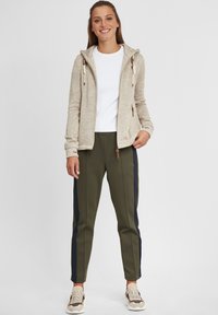 Light beige hoodie with zipper, drawstrings, and brown accents; white t-shirt; olive green tapered pants with navy side stripes; beige sneakers.