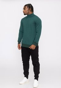 Pull à col roulé texturé vert avec des manches longues, associé à un pantalon cargo noir et des baskets blanches, présenté sur un fond neutre.