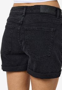 Vue arrière d'une personne portant un short en denim noir avec ourlet roulé, deux poches arrière et une étiquette en cuir de marque sur la taille.