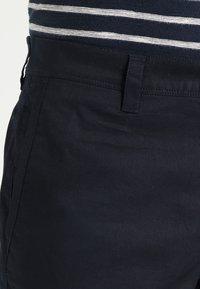 Mörkblå bomullsbyxor med en slät yta. Har en knappstängning fram och sidofickor. Randigt tyg syns ovanför midjebandet.