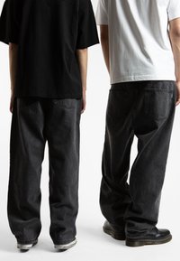 Due persone indossano jeans in denim grigio scuro a taglio largo, con tasche posteriori evidenti e stoffa testurizzata, abbinati a t-shirt nere e bianche.