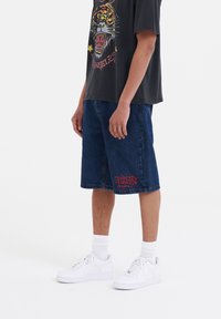 Denim shorts in dark blue with a relaxed fit and side pockets, featuring red lettering on the left leg. Paired with a black graphic tee and white sneakers.