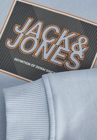 Svetlo modra dukserica z teksturirano črno in oranžno nalepko z logotipom, ki vsebuje napis "JACK & JONES" in "DEFINITION OF DENIM SINCE." Ribani manšeti.