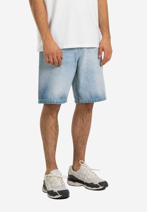 Mann trägt hellblaue Jeansshorts, weiße Turnschuhe mit schwarzen Sohlen und ein weißes T-Shirt, steht vor einem weißen Hintergrund.