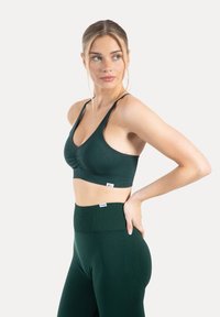 Donkergroene sportbeha met een V-hals, geplooide voorkant en verstelbare bandjes, gecombineerd met bijpassende, hooggetailleerde leggings met een gestructureerd patroon.