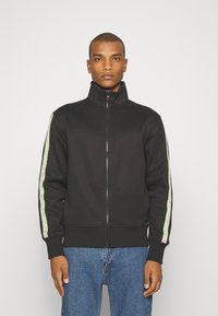 Giacca nera con zip e colletto alto, polsini e vita a coste. Presenta strisce con logo verde sui maniche. Tessuto dalla texture liscia.