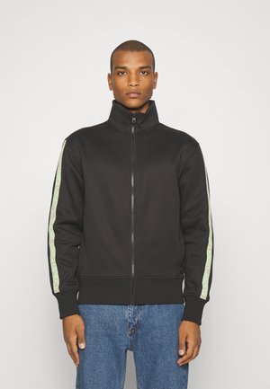 Zwarte zip-up jas met hoge kraag, ribbelmanchetten en elastische tailleband. Bevat groene logostrepen op de mouwen. Soepele stofstructuur.