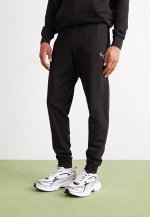 Mann trägt schwarze Puma Jogginghosen und weiße Puma Sneaker und steht auf einem grünen Boden vor weißem Hintergrund.
