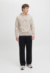Beige Kapuzenpullover mit einer Fronttasche und schwarzem Logo, kombiniert mit lockeren schwarzen Hosen und cremefarbenen Sneakern mit grauen Akzenten.