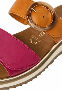 Sandale multicolore avec une lanière en cuir fuchsia et une bande de cheville en cuir couleur tan, ornée d'un accent circulaire marron et d'une semelle intérieure texturée.