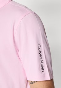 Šviesiai rožinė polo marškinėliai su trumpomis rankovėmis, tekstūruota medžiaga, su juodu "Calvin Klein" logotipu ant rankovės. Klasikinio apykaklės dizainas.