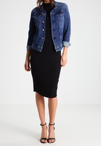 Jeansjacka med uppvikta ärmar över en figursydd svart midiklänning, tillsammans med svarta remsandaler. Outfiten har en klassisk design.