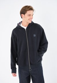 Sudadera con cremallera negra fabricada en tejido suave, con capucha forrada con patrón de cuadros, dos bolsillos frontales y un pequeño parche con el logo en el pecho.