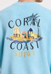 Felpa blu chiaro con una grafica di capanne da spiaggia e palme, con il testo "CORAL COAST SHIW" in grassetto nero e giallo.