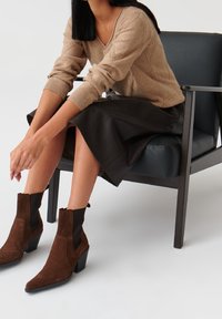 Bottines en suède marron avec des panneaux élastiques noirs sur les côtés et un talon épais, associées à un pull en laine beige et une jupe en cuir noire.