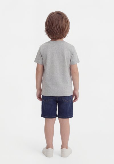 Barn står med ryggen mot, iført en grå kortermet skjorte, mørke denimbukser og hvite joggesko på hvit bakgrunn.