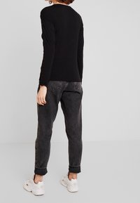 Schwarzes Langarmshirt mit tailliertem Design und Knopfakzenten an den Ärmeln, kombiniert mit locker sitzenden, dunklen Jeans und weißen Sneakers.