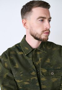 Veste verte à motif camouflage en tissu texturé, avec un devant à boutons et une poche poitrine à deux boutons.