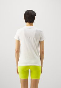 Hvid bomulds t-shirt med korte ærmer, parret med tætsiddende neongule shorts, set fra ryggen mod en ensfarvet baggrund.