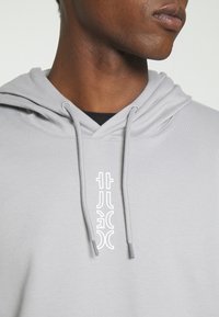 Gros plan d'une personne portant un sweat à capuche gris clair avec un texte de logo vertical blanc et des cordons visibles sur la poitrine.