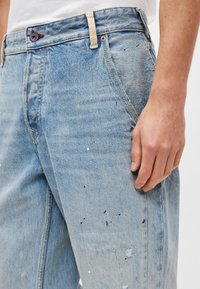 Μπλε τζιν παντελόνι με ξεβαμμένο πλύσιμο, διακοσμημένο με πιτσιλιές χρώματος, φθαρμένες λεπτομέρειες στο τελείωμα και κλασικό σχέδιο με πέντε τσέπες.