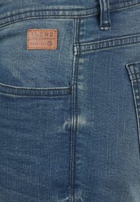 Jeans iz denima v opranem modrem odtenku z teksturirano površino. Imajo usnjeno oznako z napisom "BLEND" blizu zadnjega žepa, vidno šivanje.