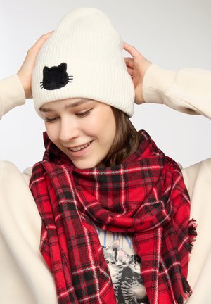 Biała, prążkowana czapka z detalem czarnej kociej twarzy oraz czerwona szalik w kratę z frędzlami, noszona na jasnym swetrze.