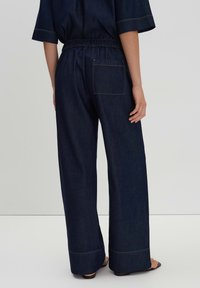 Denim wide-leg pants with an elastic waistband, dark blue color, single back pocket, and contrasting yellow stitching detail.