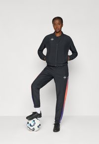 Μαύρο αθλητικό μπουφάν με φερμουάρ μπροστά, συνδυασμένο με μαύρο παντελόνι που έχει έντονες πορτοκαλί και μωβ κάθετες λωρίδες. Στέκεται πάνω σε μια μπάλα ποδοσφαίρου.