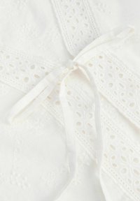 Tissu blanc avec broderie ajourée et un détail de nœud noué réalisé à partir de bandes de tissu assorties.