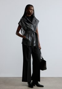 Zwarte leren mouwloze jas met een tailleband, brede revers en drukknopen, gecombineerd met zwarte wijde broekspijpen en enkellaarsjes.