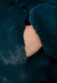 Manteau en fausse fourrure teal avec une surface douce et texturée. Une main est positionnée contre le tissu près de l'ouverture circulaire de la manche.