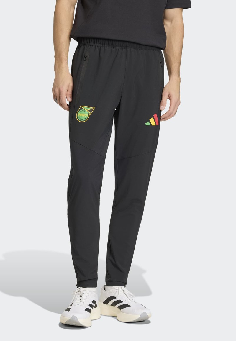 Uomo con pantaloni sportivi neri con i loghi della Jamaica Football Federation e Adidas, abbinati a sneakers bianche Adidas con strisce nere.