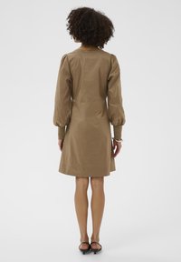 Göndör hajú nő, aki háttal áll, térd hosszúságú barna ruhát visel hosszú puffos ujjal és fekete kis sarkú cipővel.