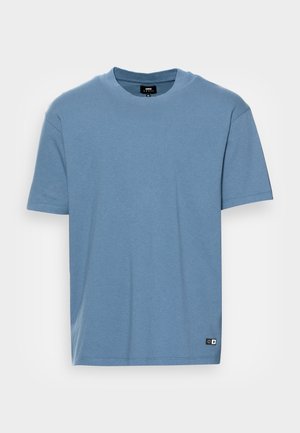 T-shirt à manches courtes en coton bleu clair, col rond, avec une petite étiquette logo en bas à gauche. Texture lisse, coupe régulière.