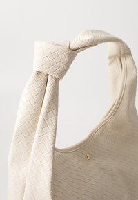 Sac à main tissé beige avec une finition texturée, présentant une bandoulière nouée et un subtil accent doré. Matériau lisse et doux.